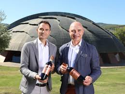 Two men holding wine bottles in front of a large arched winery building