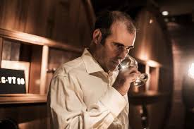 Man tasting from a glass in a winery, wearing a light shirt; barrels nearby