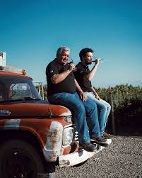 Two people posing with wine glasses outdoors beside a pickup truck